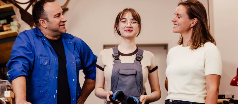 Drei Personen stehen in einer Holzwerkstatt. In der Mitte steht eine Jugendliche mit Arbeitskleiung und blickt selbstbewusst in die Kamera. Rechts und links stehen ihr zugewandt lächelnd ein Mann in Arbeitskleidung und eine Frau in Jeans und TShirt.