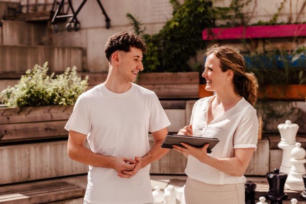 Beratungssituation in einem Innenhof: eine Beraterin erklärt einem Jugendlichen etwas an einem Tablet;