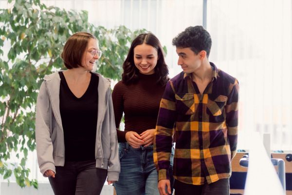 Drei Jugendliche gehend in einem Gang; sie schauen sich gegnseitig an und lächeln;