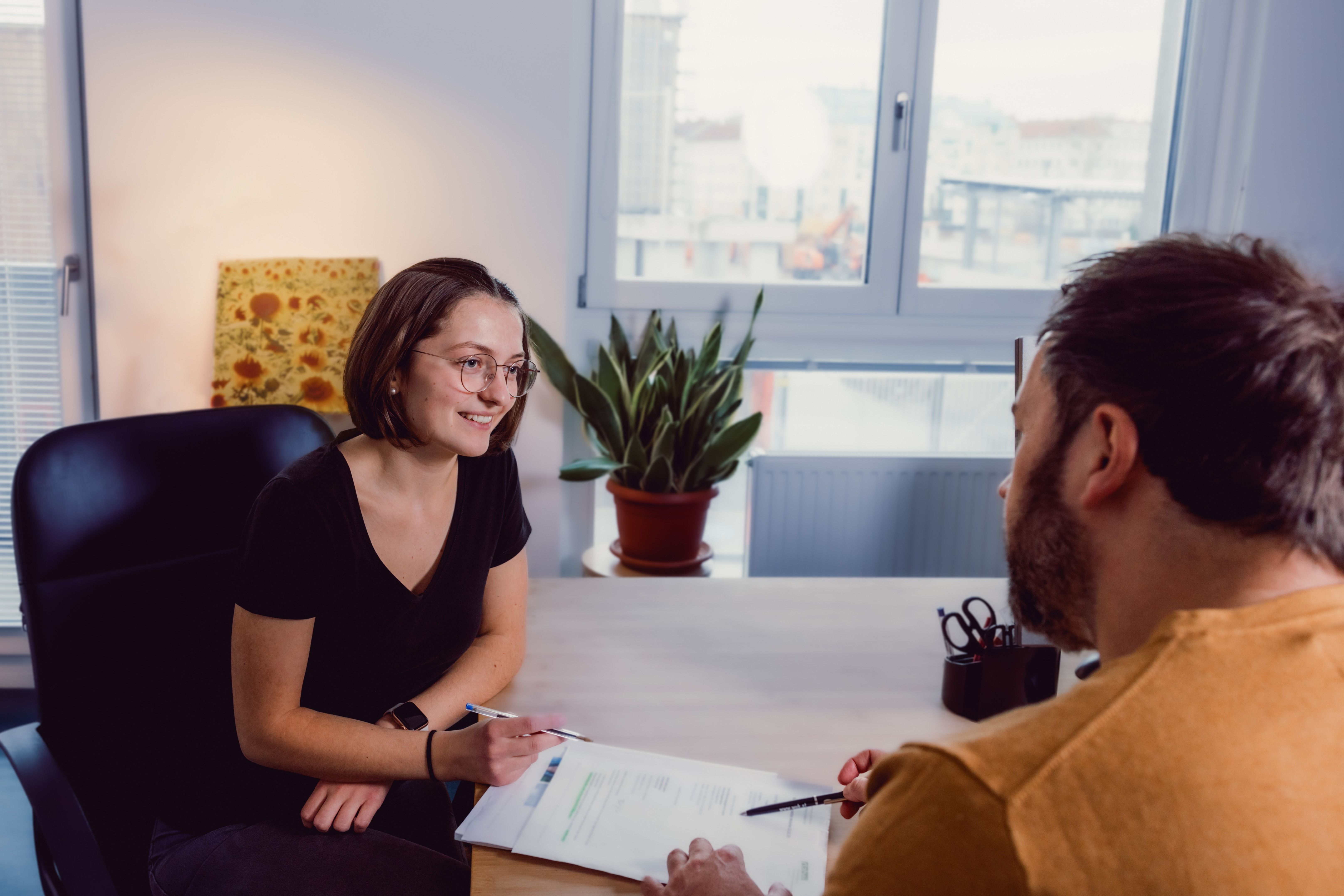Beratungssituation: eine junge Frau sitzt lächelnd gegenüber einem Betreuer in einem Büro. Auf dem Schreibtisch liegen Unterlagen und im Hintergrund ist ein Bild mit Sonnenblumen zu sehen.